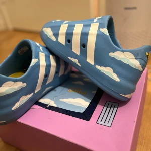 Blå adiFOM Superstar Simpsons från Adidas - Unika blå adiFOM Superstar sneakers från Adidas i samarbete med The Simpsons. Skorna har ett lekfullt molnmönster och de ikoniska tre ränderna i vitt. Perfekta för den som vill sticka ut med en touch av humor och stil.