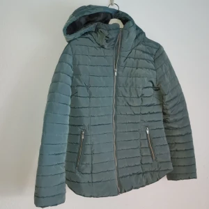 Grön dunjacka från Lindex - Säljer en snygg grön dunjacka från Lindex. Jackan har en dragkedja framtill och två praktiska fickor med dragkedjor. Den är quiltad och har en huva för extra värme. Perfekt för kyliga dagar!