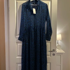 Blå leopardmönstrad långklänning - Snygg blå långklänning med leopardmönster. Klänningen har långa ärmar och en avslappnad passform, perfekt för en stilfull look. Den är tillverkad i ett lätt och luftigt material.