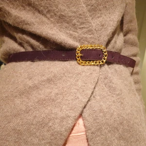 Mörklila bälte med guldfärgat spänne - Snyggt mörklila bälte med ett iögonfallande guldfärgat kedjespänne. Perfekt för att ge din outfit en extra touch av stil. Bältet är smalt och elegant, vilket gör det lätt att matcha med olika kläder. Mocka/ mockaimitation 