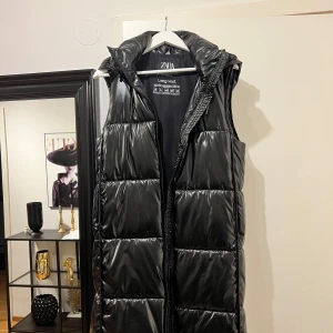 Svart lång dunväst från Zara - Snygg och praktisk svart lång dunväst från Zara. Västen har en glansig yta och är utrustad med dragkedja. Perfekt för kyligare väder med sin vattenavvisande och vindtäta design. Den har en huva för extra skydd. västen har inre och yttre fickor. 