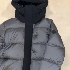 Moncler jacka - Säljer en snygg grå dunjacka från Moncler med hög krage och huva. Jackan har en stilren design med knappar och dragkedja framtill. Perfekt för kyliga dagar. Storlek M men passar till L. Helt ny, aldrig användt! 