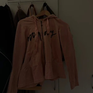 Rosa hoodie från Odd Molly - Säljer en snygg rosa hoodie från Odd Molly med dragkedja och tryck framtill. Den har en cool logga på ärmen och är perfekt för en avslappnad stil. Huvan har snören för justering. Perfekt för dig som gillar en bekväm och stilren look. Den är jätte 2000s 