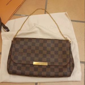 Louis Vuitton väska (Damier Ebene) - ❗️Skriv innan köp❗️ Snygg brun rutig axelväska i model Favorite MM D. EBE. från Louis Vuitton med guldfärgad kedja. Väskan har ett elegant rutmönster och en röd insida med praktiska fack. Har används men omhändertagen. Det är äkta Louis Vuitton köpt 2018 i Dubai, kvitto och låda är inkluderat. 