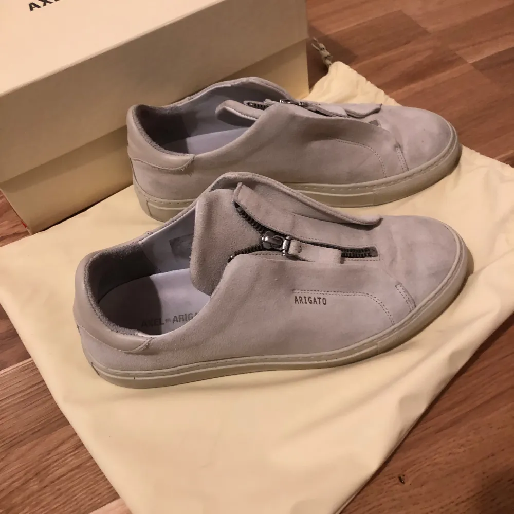 Snygga grå sneakers från Axel Arigato i mocka med dragkedja framtill. Tillverkade i Portugal och kommer med originalförpackning. Perfekta för en stilren look. Storlek 41. Kengät.