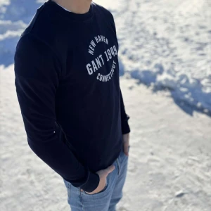 Svart långärmad tröja från GANT - Säljer en stilren svart långärmad tröja från GANT med tryck 'New Haven GANT 1949 Connecticut' på bröstet. Perfekt för en avslappnad look. Tröjan har ribbade muddar vid ärmslut och nederkant.