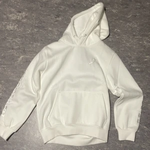 Vit Jordan hoodie med tryck - Säljer en stilren vit hoodie med tryck på ärmen och bröstet. Den har en klassisk känguruficka och justerbar huva. Perfekt för en avslappnad stil.