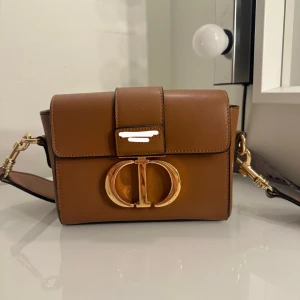 Brun axelväska med gulddetaljer - Snygg brun axelväska i skinn med guldfärgad logodetalj framtill. Väskan har en justerbar axelrem med mönster och guldfärgade spännen. Perfekt för att ge en touch av lyx till din outfit.