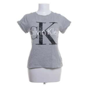 Grå t-shirt från Calvin Klein - Snygg grå t-shirt från Calvin Klein med stort tryck av logotypen på framsidan. Den har korta ärmar och en klassisk rund halsringning. Perfekt för en avslappnad stil.