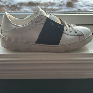 Valentino Garavani sneakers i vitt och svart - Snygga Valentino Garavani sneakers i vitt skinn med en svart detalj över snörningen. Skorna har en klassisk design med nitar på hälen och en bra sula. Perfekta för en stilren look. Inget og följer med