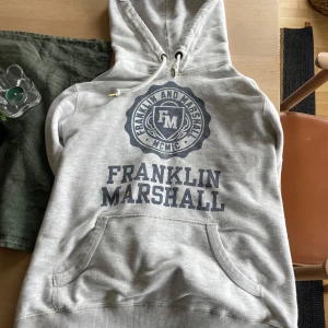 Grå hoodie från Franklin & Marshall - Säljer en grå hoodie från Franklin & Marshall med tryck framtill. Den har en klassisk känguruficka och justerbar huva med snören. Perfekt för en avslappnad stil.