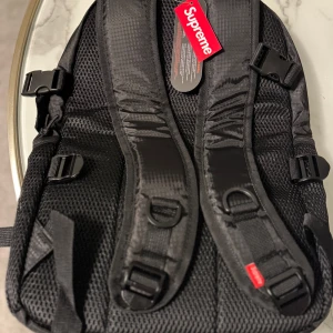 Svart ryggsäck från Supreme - Snygg svart ryggsäck från Supreme i slitstarkt Cordura-material. Väskan har justerbara axelremmar och flera spännen för extra säkerhet. Perfekt för att bära dina grejer med stil. Den har en stor huvudficka med dragkedja och Supreme-loggan i vitt på framsidan.