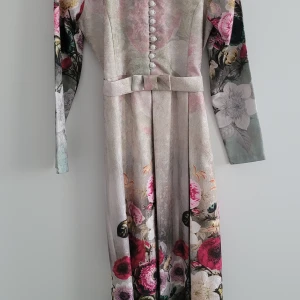 Blommig långklänning med bälte - Elegant långklänning med blommigt mönster i rosa, rött och vitt. Klänningen har långa ärmar och knappar framtill samt ett matchande bälte i midjan. Perfekt för en stilfull look.