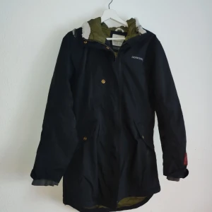 Svart parkajacka från Didriksons - Säljer en svart parkajacka från Didriksons med huva och dragkedja. Jackan har en quiltad insida och knappar framtill. Perfekt för kyligare dagar med sin långa passform och praktiska fickor.