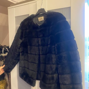 Svart pälsjacka från Estile - Snygg svart pälsjacka från Estile med en lyxig och mjuk känsla. Jackan har en klassisk design med långa ärmar och en rak passform. Perfekt för att hålla sig varm och stilren under kyliga dagar.