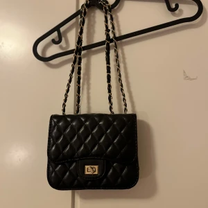 Svart quiltad axelväska - Snygg svart quiltad axelväska med kedjedetaljer på axelremmen. Väskan har en elegant låsdetalj framtill och är perfekt för att ge en stilren touch till din outfit. Den kompakta storleken gör den smidig att bära med sig.