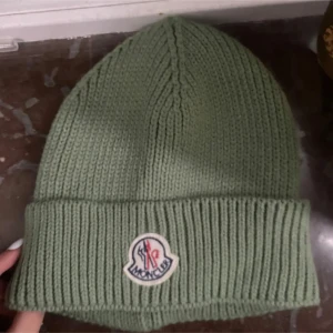 Grön stickad mössa  - Snygg grön stickad mössa från Moncler med uppvikt kant och broderad logotyp framtill. Perfekt för kyliga dagar. Ej äkta!- oanvänd 