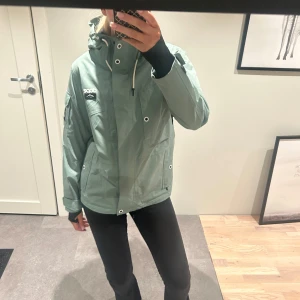 Grön skidjacka från Dope - Snygg grön skidjacka från Dope med huva och dragkedja. Jackan har långa ärmar och praktiska fickor framtill. Perfekt för blåsiga dagar och har en stilren design med vita detaljer.