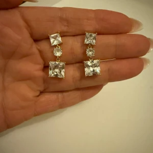 Guldörhängen med fyrkantiga stenar - Eleganta guldörhängen med två fyrkantiga klara stenar och en rund sten i mitten. Perfekta för att ge en glittrig touch till din outfit.