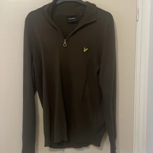 Olivgrön tröja med dragkedja från Lyle & Scott - Snygg olivgrön tröja från Lyle & Scott med dragkedja vid halsen och deras ikoniska logga på bröstet. Perfekt för en stilren och avslappnad look. Tröjan har långa ärmar och en bekväm passform.