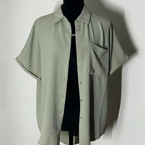Grön kortärmad skjorta - Snygg och avslappnad grön skjorta med korta ärmar och knappar framtill. Perfekt för en casual look med en bröstficka för extra stil. Skjortan har en loose passform som ger en bekväm känsla.