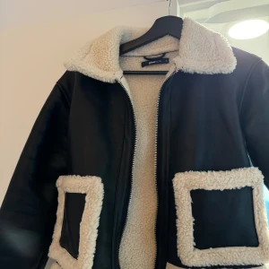 Svart jacka med teddyfoder från Gina Tricot - Säljer en snygg svart jacka från Gina Tricot med mysigt teddyfoder. Jackan har en dragkedja framtill och två stora fickor med teddykanter. Perfekt för kyliga dagar när du vill hålla dig varm och stilren.