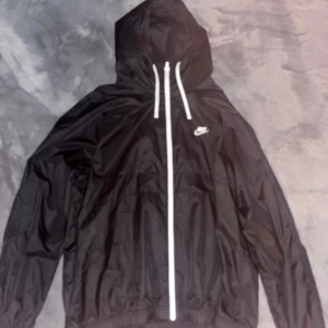 Svart vindjacka från Nike - Säljer en svart vindjacka från Nike med huva och dragkedja. Jackan har en klassisk design med Nike-logga på bröstet och justerbara snören vid huvan. Perfekt för blåsiga dagar.