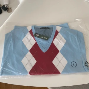 Ljusblå tröja med argylemönster från Pelican - Snygg ljusblå tröja från Pelican med klassiskt argylemönster i rött och vitt. Tröjan har en v-ringad hals och är perfekt för en stilren look. Den är förpackad i plast och i helt nytt skick.