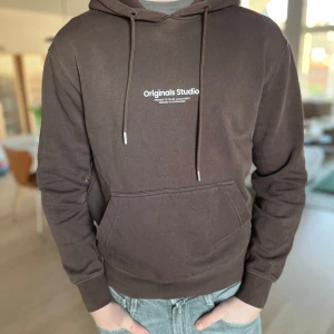Brun Jack&jones hoodie  - Säljer en snygg brun hoodie från Originals Studio med vit text på bröstet. Den har en klassisk känguruficka och justerbar dragsko i huvan. Perfekt för en avslappnad stil. Ny pris 799kr nu endast 350kr