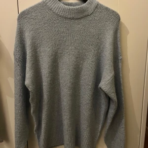 Ljusblå stickad tröja från 157 - Mysig ljusblå stickad tröja från 157 med en hög krage. Perfekt för kyliga dagar med sin mjuka och bekväma passform. Tröjan har långa ärmar och ribbade detaljer vid krage, ärmslut och nederkant. Använd ca 3-5 gånger