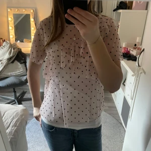 Rosa prickig blus med volang - Säljer en söt rosa blus med svarta prickar och volangdetaljer. Blusen har korta ärmar och är gjord i ett lätt och luftigt material som även är ganska genomskinligt. Den är i storlek L men sitter som en storlek S/M.