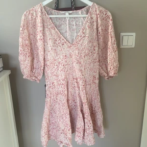 Rosa och vit klänning från H&M - Säljer en söt rosa och vit klänning från H&M med ett abstrakt mönster. Klänningen har en v-ringad hals och korta puffärmar. Perfekt för varma dagar med sitt luftiga material och avslappnade passform.