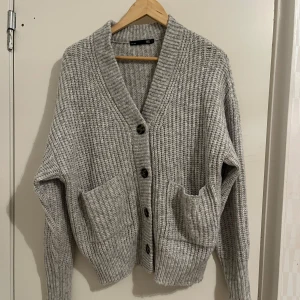 Grå stickad kofta från 157 - Mysig grå stickad kofta från 157 med stora knappar och två praktiska fickor framtill. Perfekt för kyliga dagar med sin ribbade struktur och v-ringad design. Ett måste i garderoben för en stilren och bekväm look.