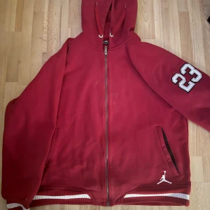 Röd hoodie från Jordan - Säljer en röd hoodie från Jordan med dragkedja och huva. Den har ett stort '23' tryck på ärmen och den ikoniska Jumpman-loggan på framsidan. Perfekt för en sportig look.