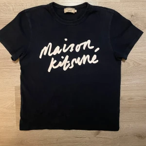 Svart t-shirt från Maison Kitsuné - Är size small, men ganska liten i storlek. Snygg svart t-shirt från Maison Kitsuné med vit text på framsidan. Perfekt för en stilren och avslappnad look. Gjord i mjukt material för extra komfort.