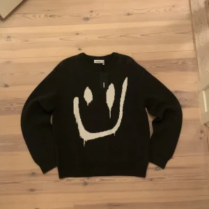 Svart tröja med smiley från Weekday - Säljer en svart tröja från Weekday med ett stort vitt smiley-motiv på framsidan. Tröjan har långa ärmar och en rund halsringning. Perfekt för en avslappnad stil. Priset kan såklart diskuteras vill bara bli av med den så snabbt som möjligt 