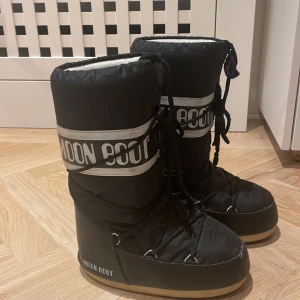 Svarta Moon Boots med snörning - Snygga och varma svarta Moon Boots med vit logga och snörning. Perfekta för kalla vinterdagar med sin robusta design och bekväma passform.