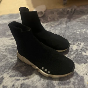Svarta sneakers med vit sula - Snygga svarta sneakers med en hög sockliknande design och vit sula. Perfekta för en stilren och modern look. De har en elastisk passform som gör dem bekväma att bära.