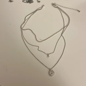 Silverfärgat halsband med tre hängen - Snyggt silverfärgat halsband med tre kedjor. Ena kedjan har ett runt hänge med ett dekorativt mönster, medan den andra har en enkel, glittrig sten. Perfekt för att lägga till lite bling till din outfit!
