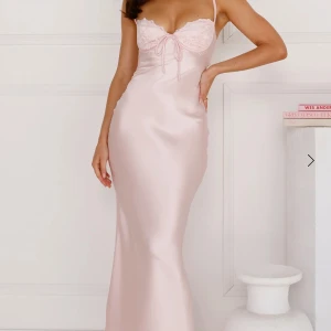 Ljusrosa långklänning i satin - Helt ny, superfin ljusrosa långklänning i satin med smala axelband och en vacker öppen rygg. Klänningen har en feminin passform och en söt knytdetalj framtill. Perfekt för speciella tillfällen som exempelvis studentbal