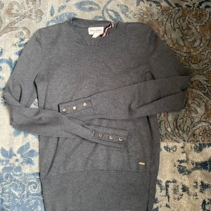 Grå tröja från Holly & Whyte - Snygg grå tröja från Holly & Whyte med rund hals och långa ärmar. Tröjan har dekorativa knappar vid ärmsluten och en diskret logga vid nederkanten. Perfekt för en stilren look.