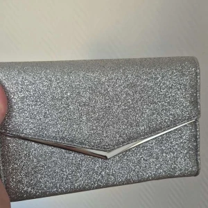 Glittrig silver kuvertväska - Snygg och glittrig kuvertväska i silver perfekt för en kväll ute. Väskan har en elegant design med en metallisk detalj på framsidan. Perfekt för att bära det nödvändigaste med stil.