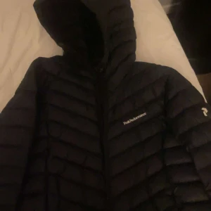 Svart dunjacka från Peak Performance - Säljer en svart dunjacka från Peak Performance med huva och dragkedja. Jackan har ett quiltat mönster och är perfekt för kyliga dagar. Den är både stilren och funktionell.