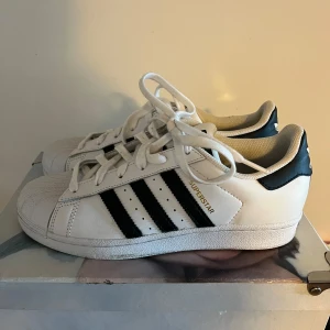Adidas Superstar i vitt och svart - Klassiska Adidas Superstar sneakers i vitt med svarta ränder och gulddetaljer. Skorna har snörning och den ikoniska tåhättan i gummi. Perfekta för en stilren och sportig look.