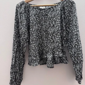 Svart blommig blus med volang - Säljer en söt svart blus med vita blommor och volangdetaljer. Blusen har långa ärmar och en luftig passform, perfekt för både vardag och speciella tillfällen. Den är gjord i ett lätt och bekvämt material.