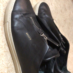 Svarta Axel Arigato - Snygga svarta sneakers från  Arigato. dragkedja på ovansidan. Skorna har en vit sula som ger en stilren kontrast. Perfekta för en avslappnad look. 