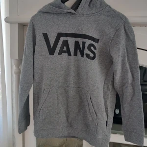 Grå hoodie från Vans - Säljer en grå hoodie från Vans med stor logga på bröstet. Tröjan har en klassisk känguruficka och ribbade muddar vid ärmslut och nederkant. Perfekt för en avslappnad stil.