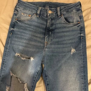 Blå jeans med slitningar - Snygga blå jeans med slitningar och en klassisk femficksdesign. De har en knapp och dragkedja framtill. Perfekta för en avslappnad stil med en modern touch.