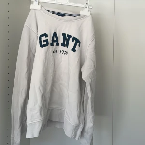 Vit sweatshirt från GANT - Säljer en vit sweatshirt från GANT med blå text 'GANT Est. 1949' på framsidan. Tröjan har långa ärmar och ribbade muddar vid ärmslut och nederkant. Perfekt för en avslappnad stil.