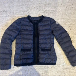 Blå dunjacka från Moncler - Säljer en snygg blå dunjacka från Moncler. Jackan har en quiltad design med långa ärmar och en rund halsringning. Perfekt för kyligare dagar och ger en stilren look. Moncler-loggan är broderad på ärmen.